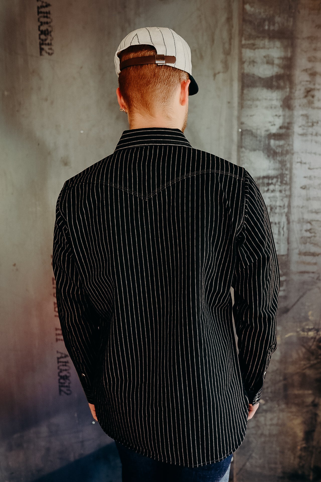 Man wearing black pinstripe shirt and white cap facing concrete wall in industrial setting