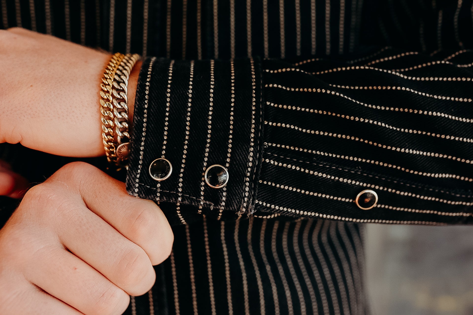 Person wearing white and gold chain bracelets with a black pinstripe shirt cuff featuring metal snap buttons