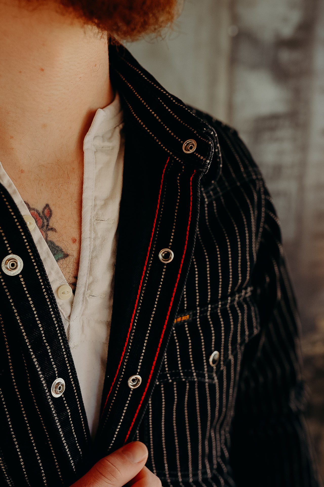 Close-up of man wearing black pinstripe jacket with metal snap buttons over white shirt, chest tattoo visible