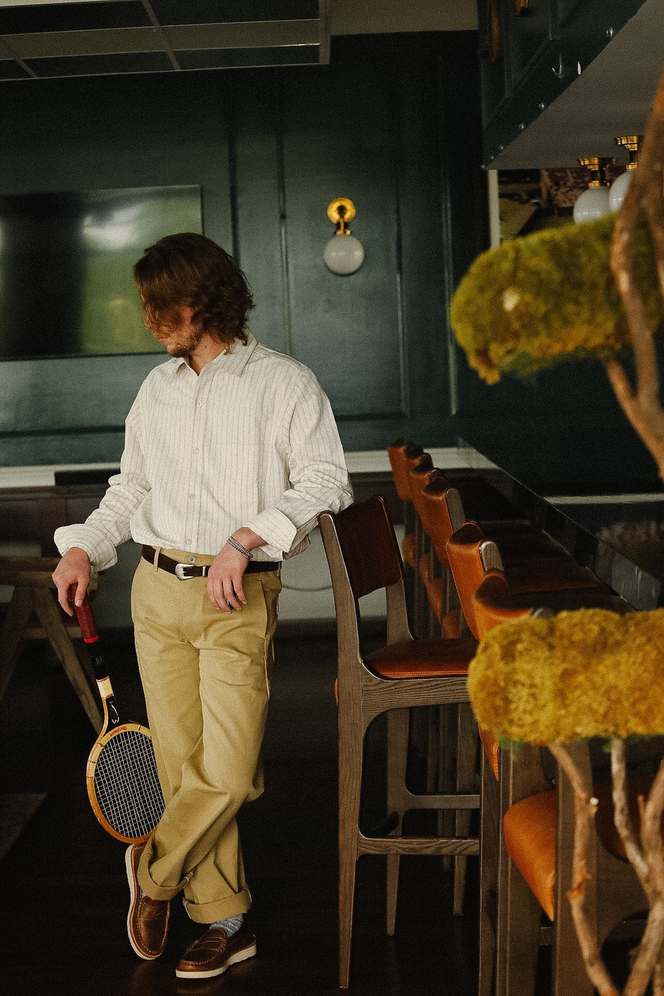Man in a white shirt and beige pants standing in a bar with a badminton racket, surrounded by bar stools and a green wall.
