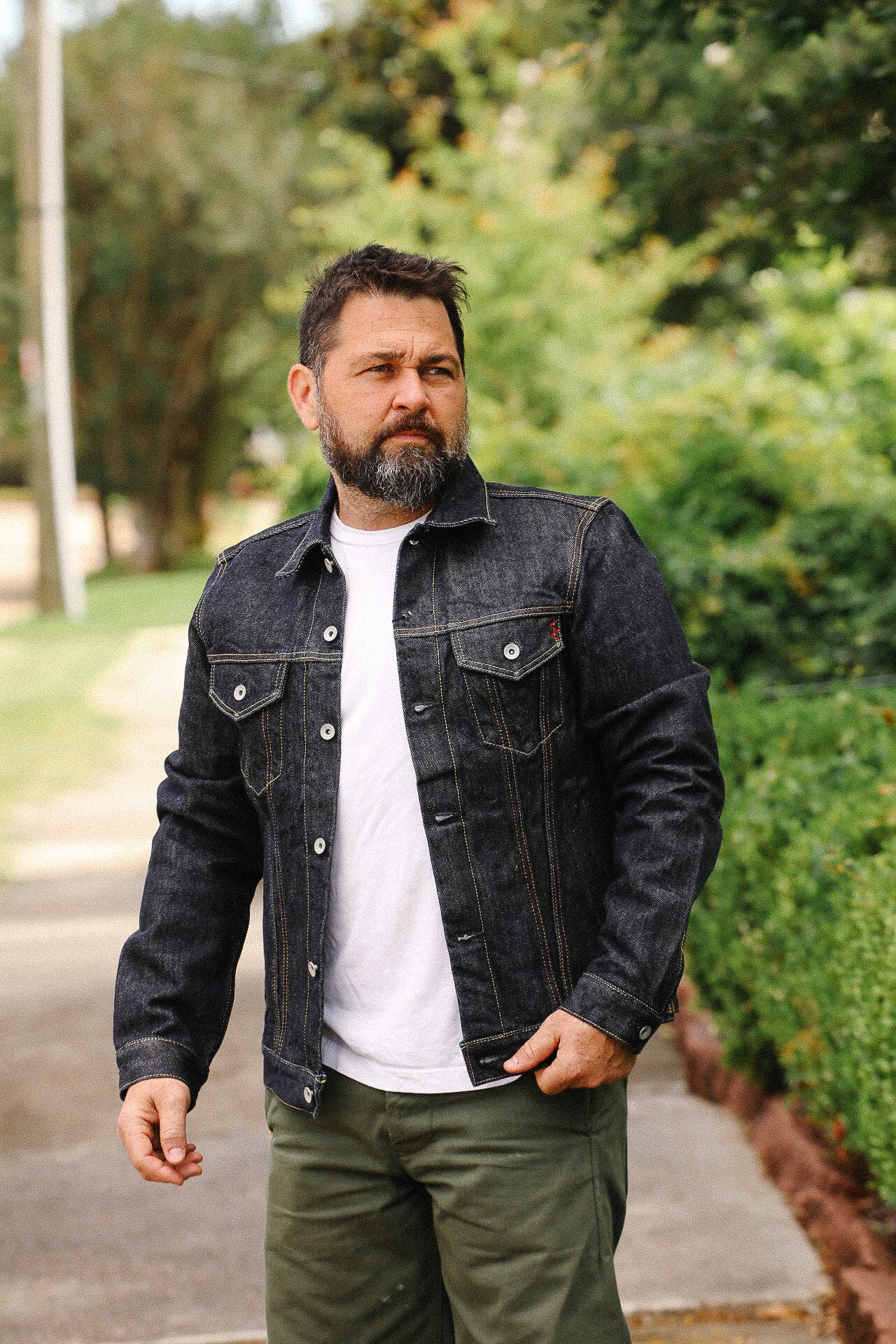 Bearded man wearing black denim jacket and white t-shirt walking outdoors near greenery