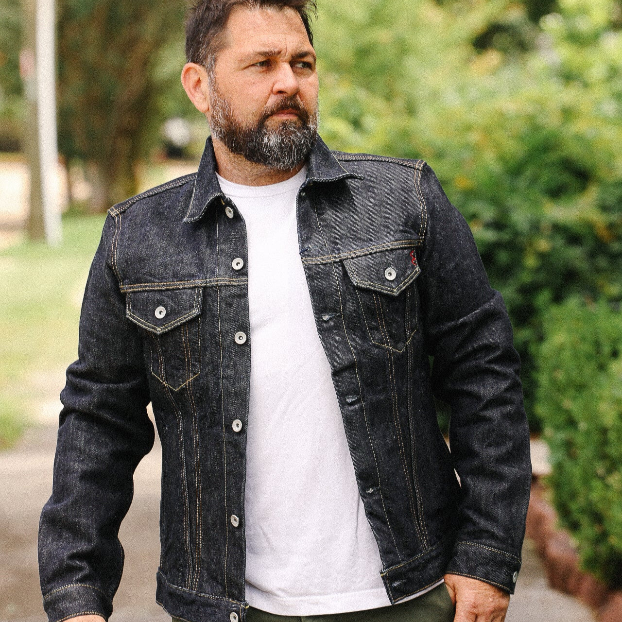 Bearded man wearing a dark denim jacket and white shirt standing outdoors near green bushes