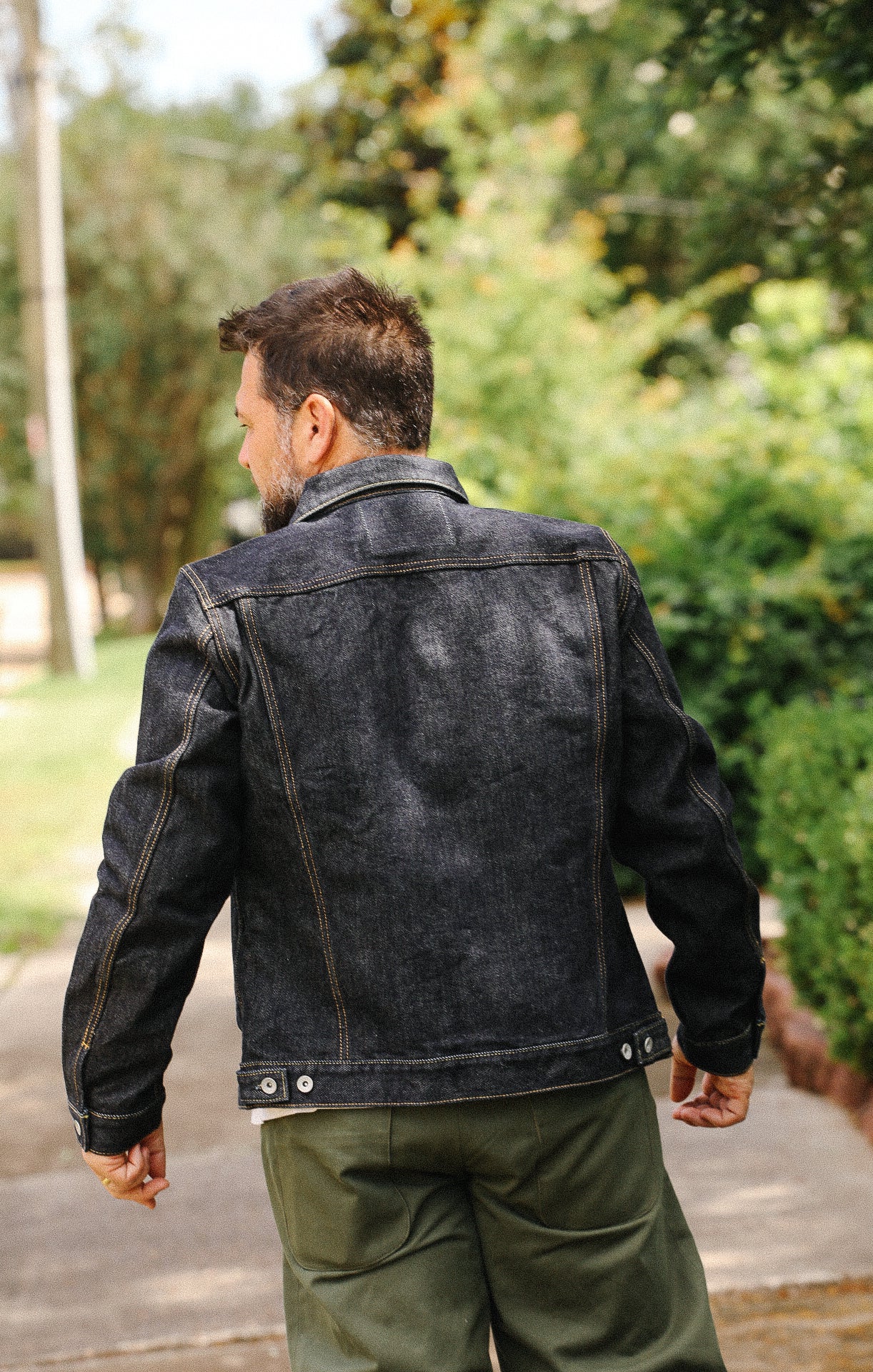 Man wearing dark denim jacket and olive green pants walking outdoors on a sunny day