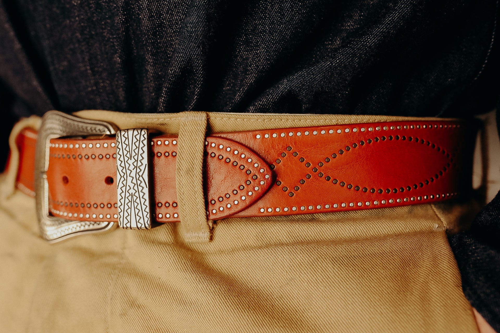 Close-up of brown leather belt with metal studs and engraved silver buckle worn with tan pants and dark denim shirt