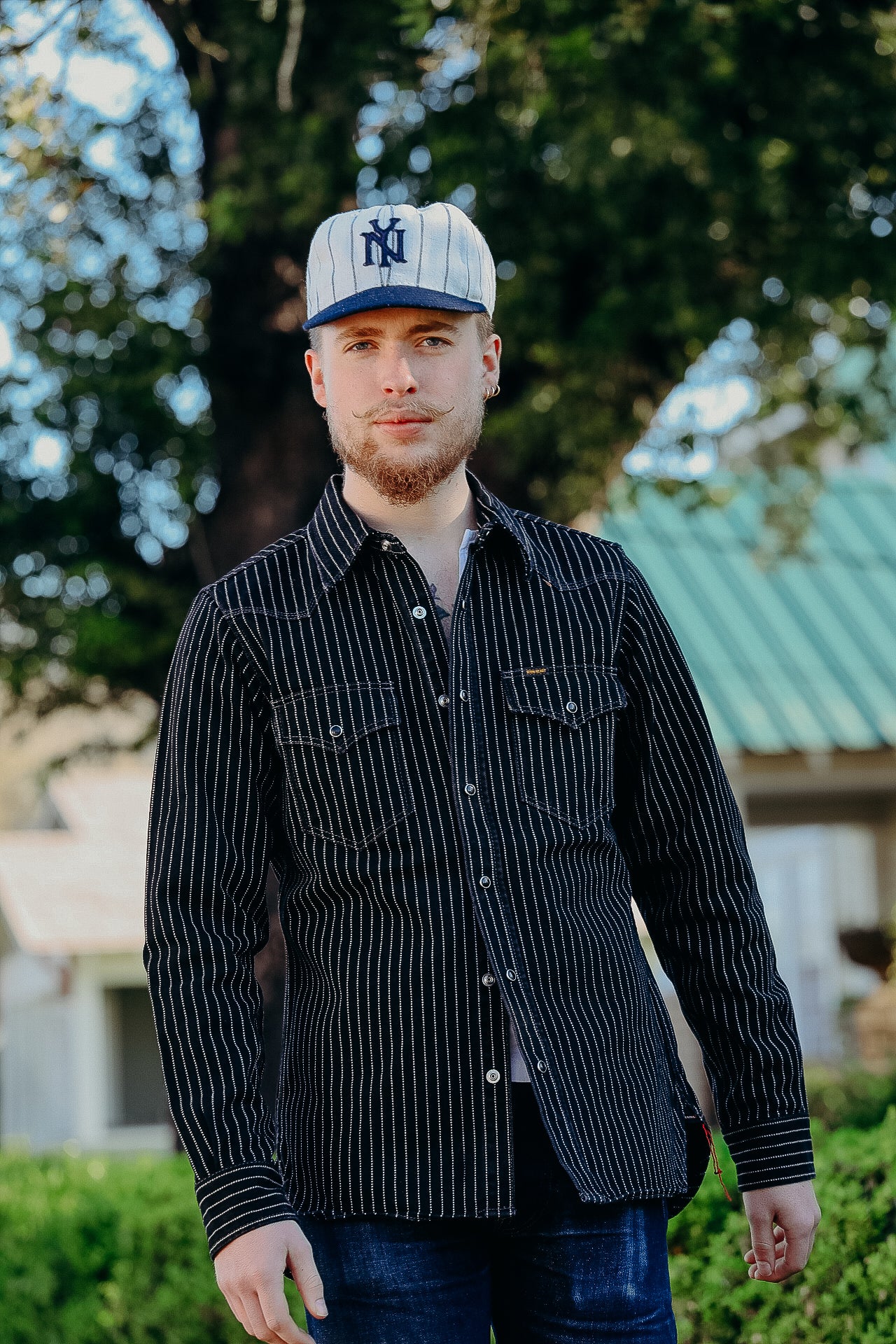 Man wearing a black pinstripe button-up shirt and vintage style cap outdoors with greenery background