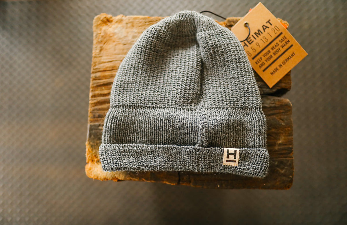 Gray knit beanie hat with a leather Heimating tag, displayed on rustic wooden surface