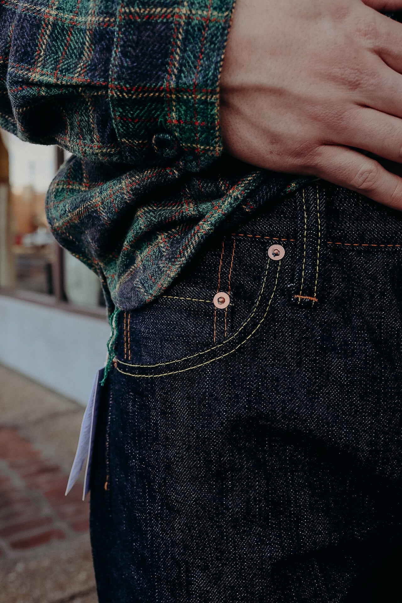 Close-up of hand on dark denim jeans pocket with detailed yellow and orange stitching, worn with green plaid shirt