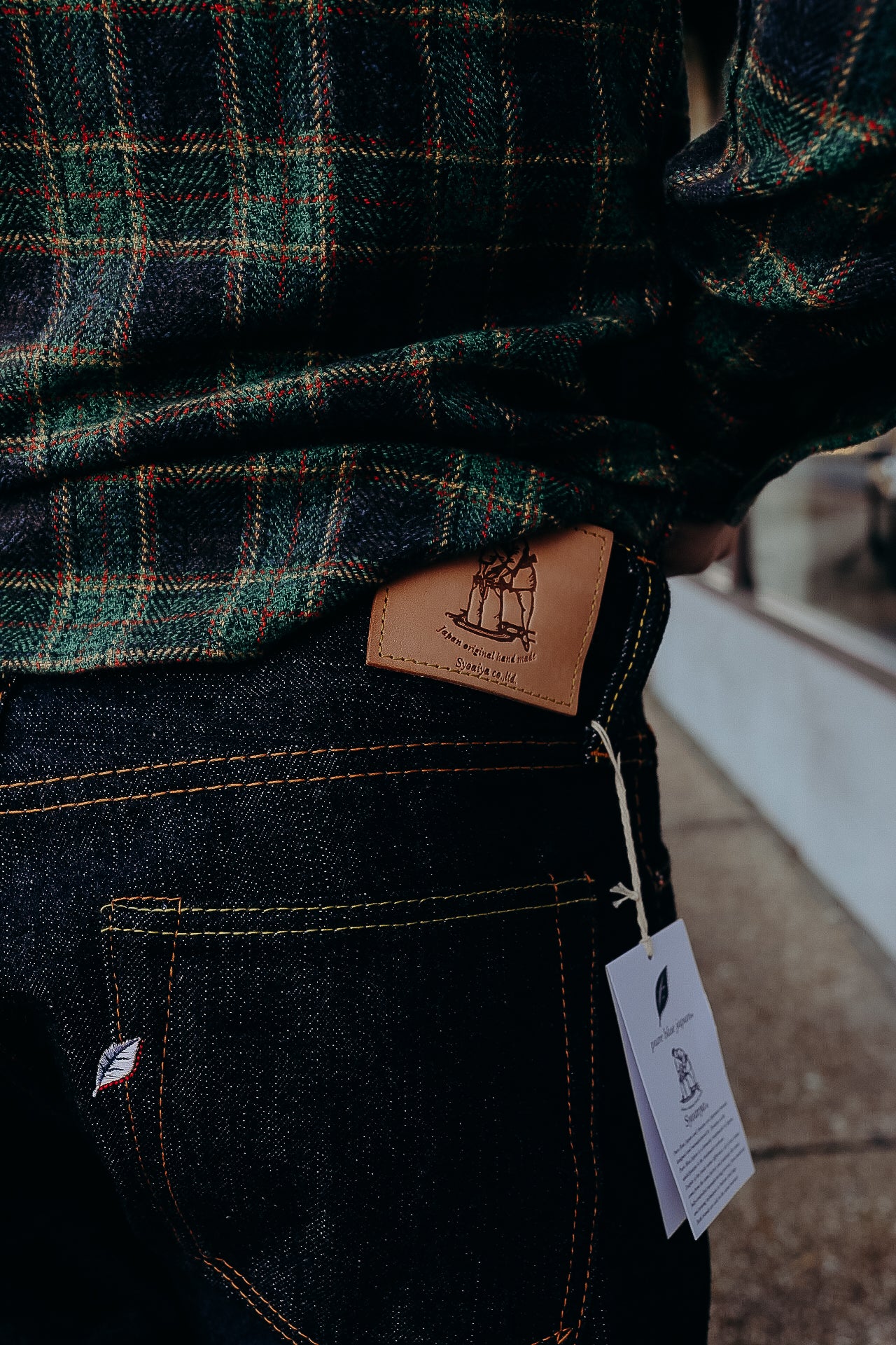 Close-up of dark denim jeans with leather patch and hang tag, paired with green plaid shirt