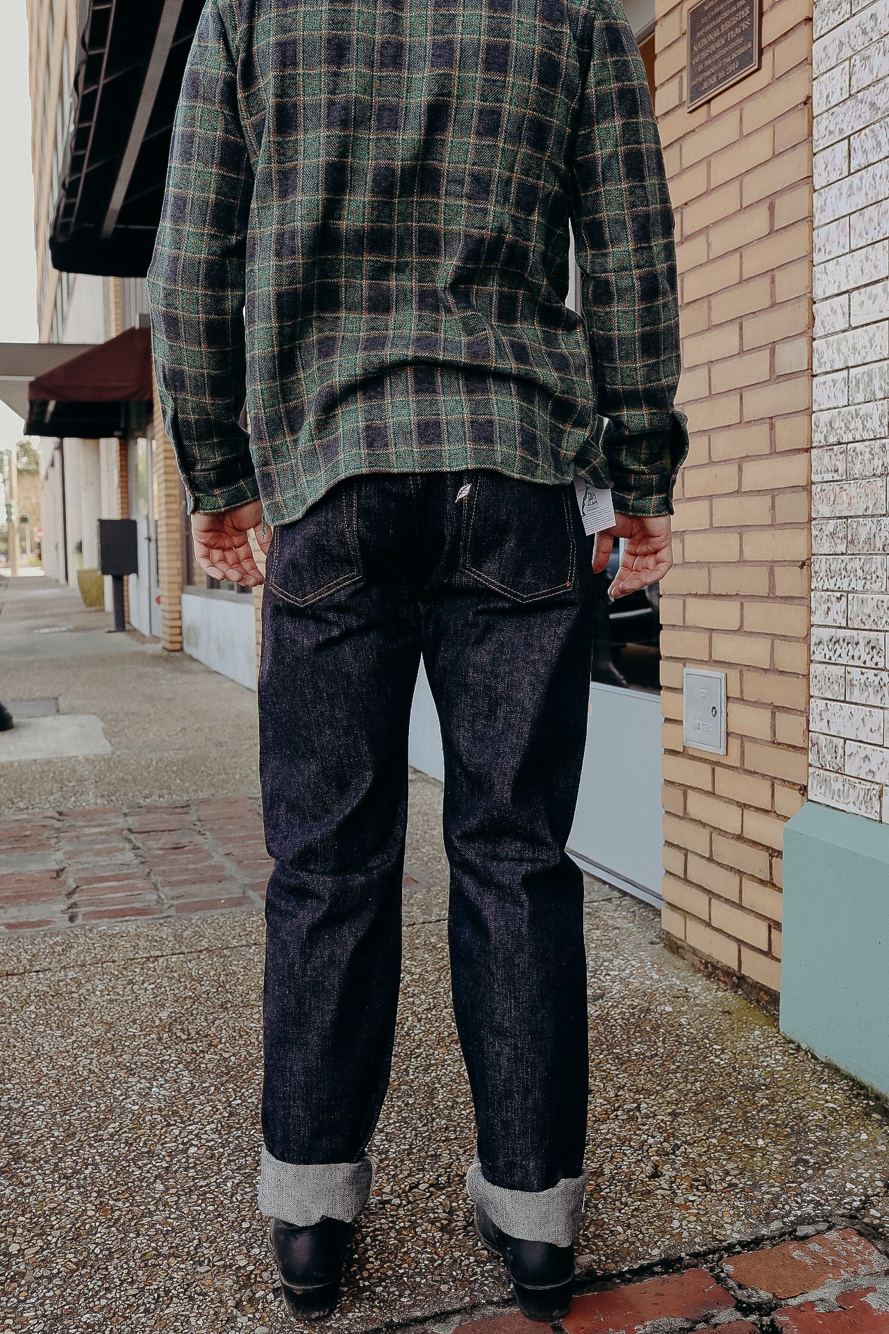Person wearing dark raw denim jeans cuffed at the bottom and a green plaid shirt standing on a city sidewalk