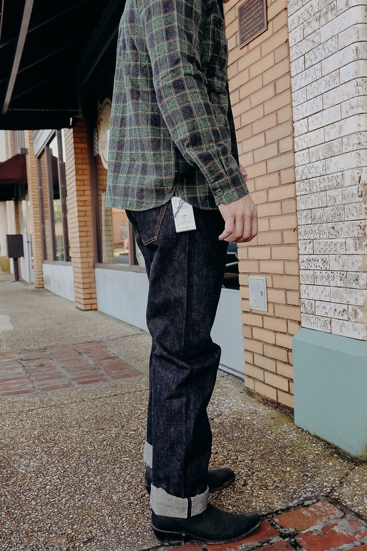 Side view of a person standing outside a brick building wearing dark selvedge denim jeans with cuffs, black boots, and a green plaid shirt
