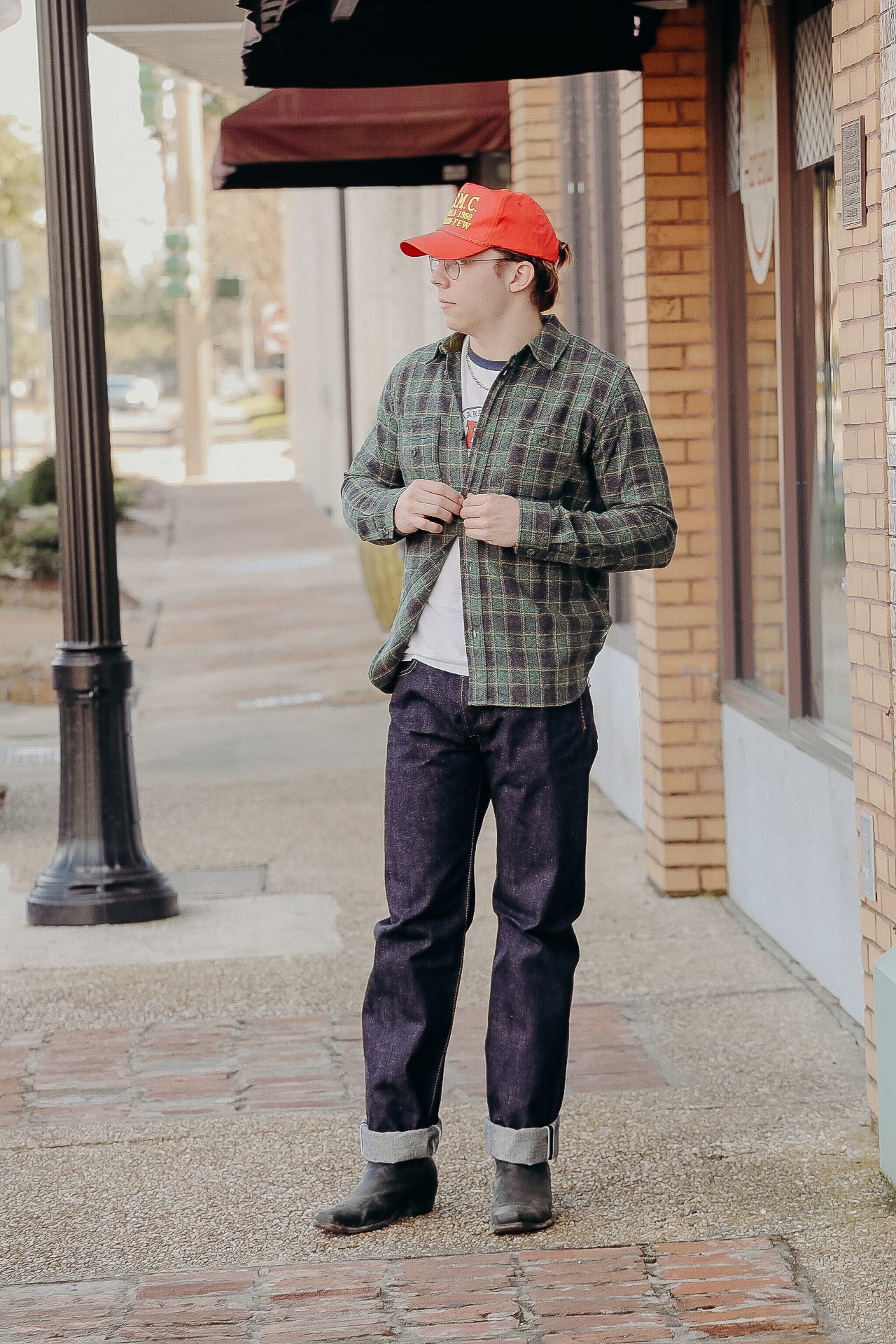 Man wearing red baseball cap, glasses, green plaid shirt, cuffed dark jeans, and boots standing on sidewalk