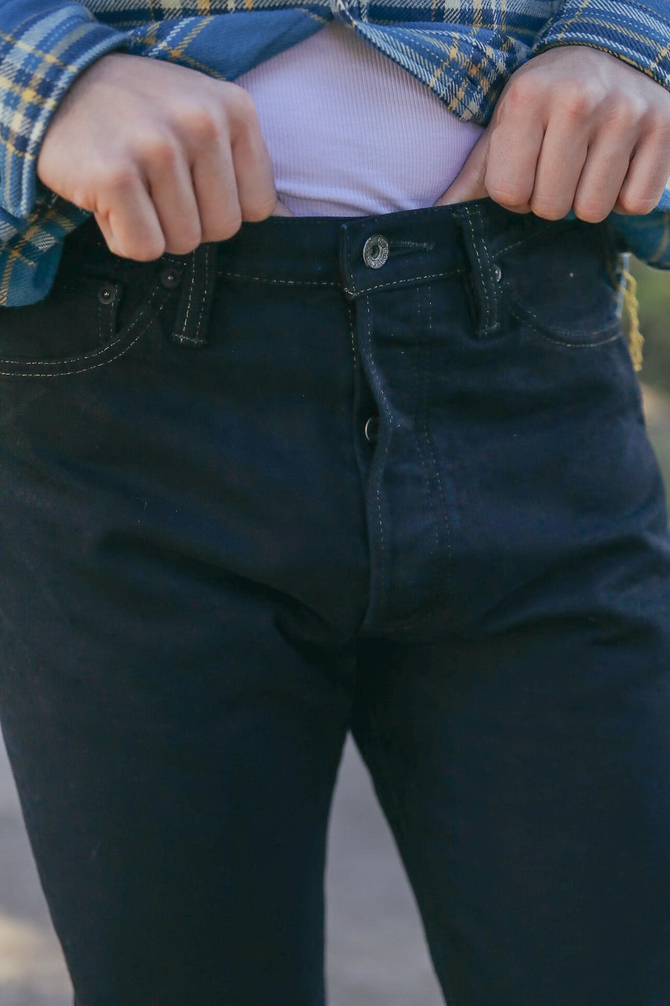 Close-up of person fastening button on black denim jeans wearing blue plaid shirt