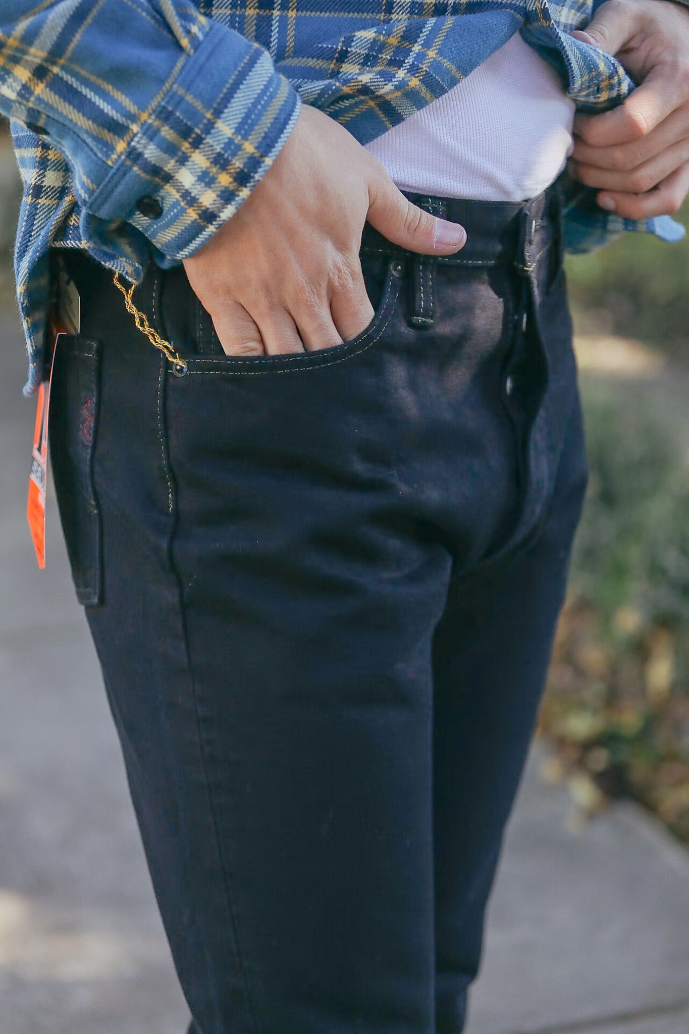 Person wearing black rugged work pants with hands in pockets and a blue plaid shirt outdoors