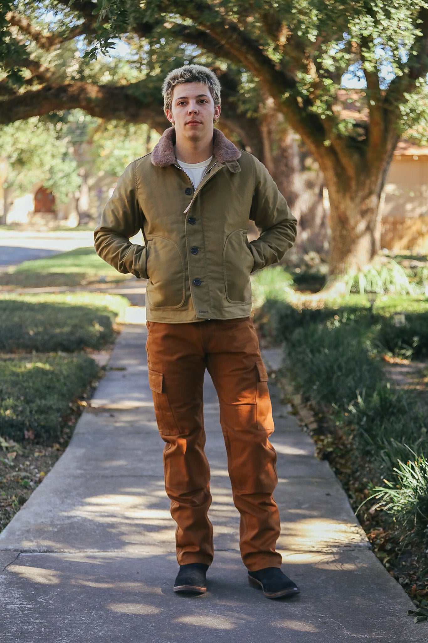 Man wearing green jacket with shearling collar and brown cargo pants standing on sidewalk amid trees