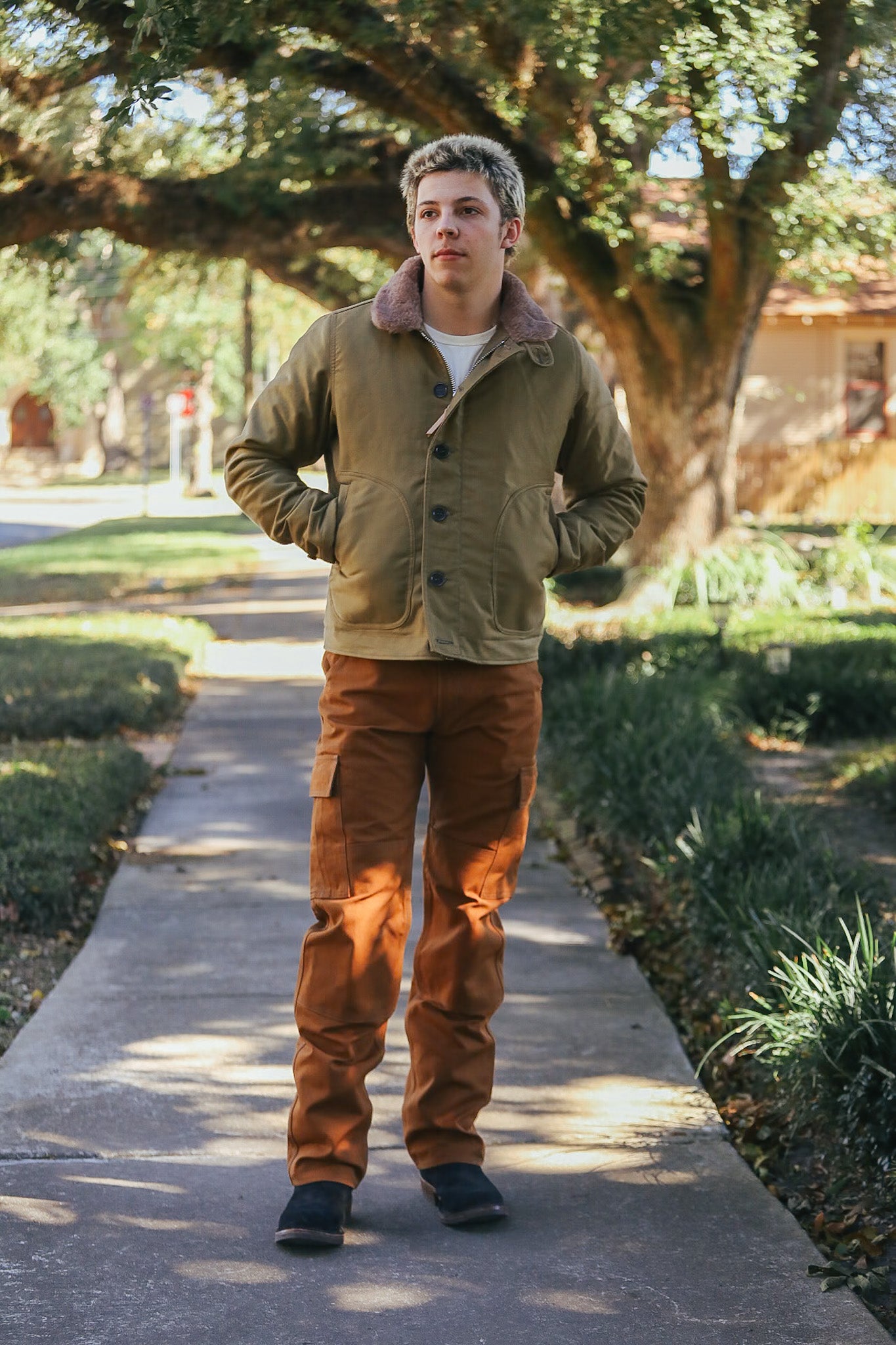 Man wearing olive canvas jacket with shearling collar and brown cargo pants on suburban sidewalk