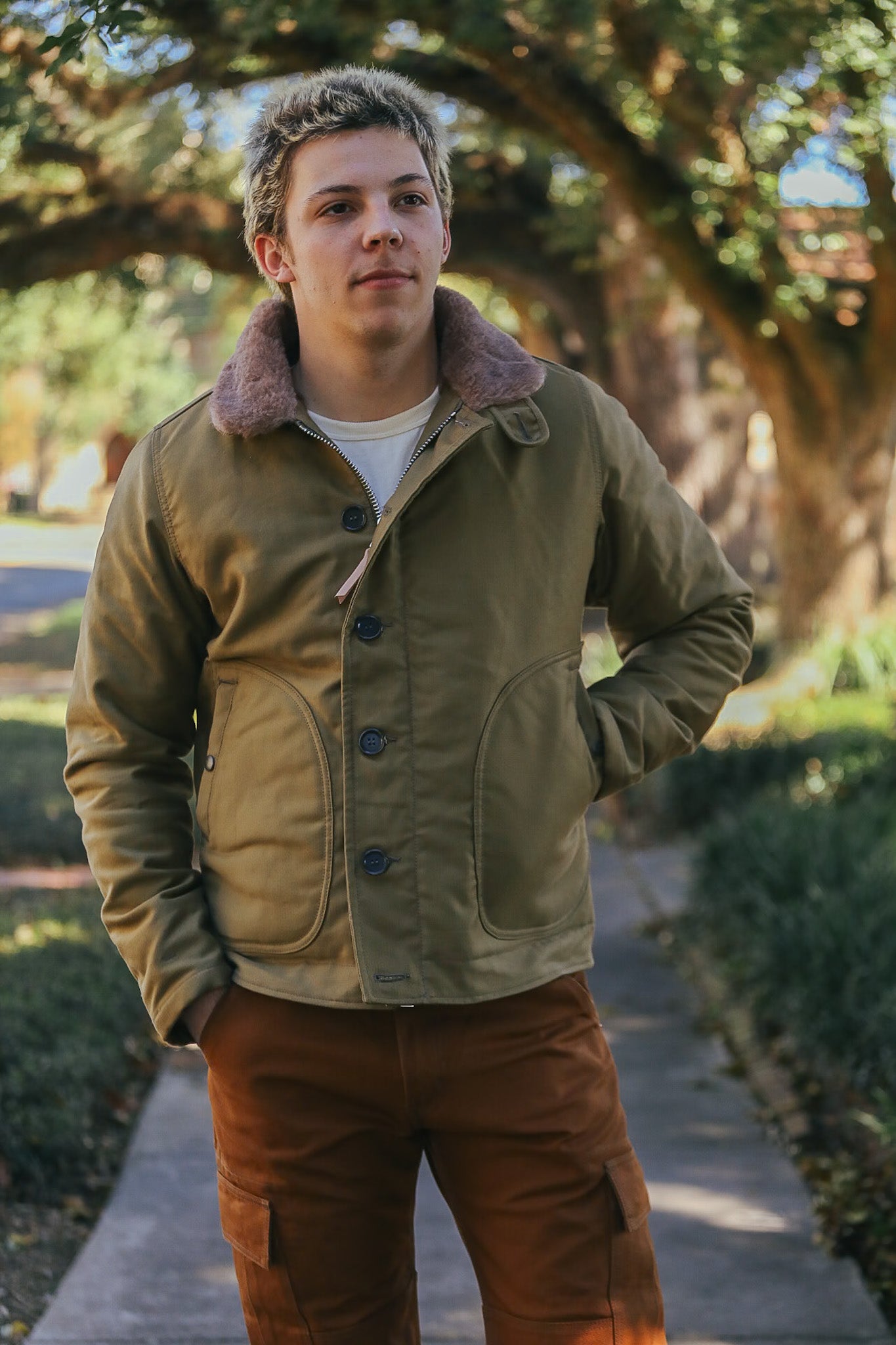 young man wearing olive green buttoned jacket with shearling collar and brown cargo pants outdoors