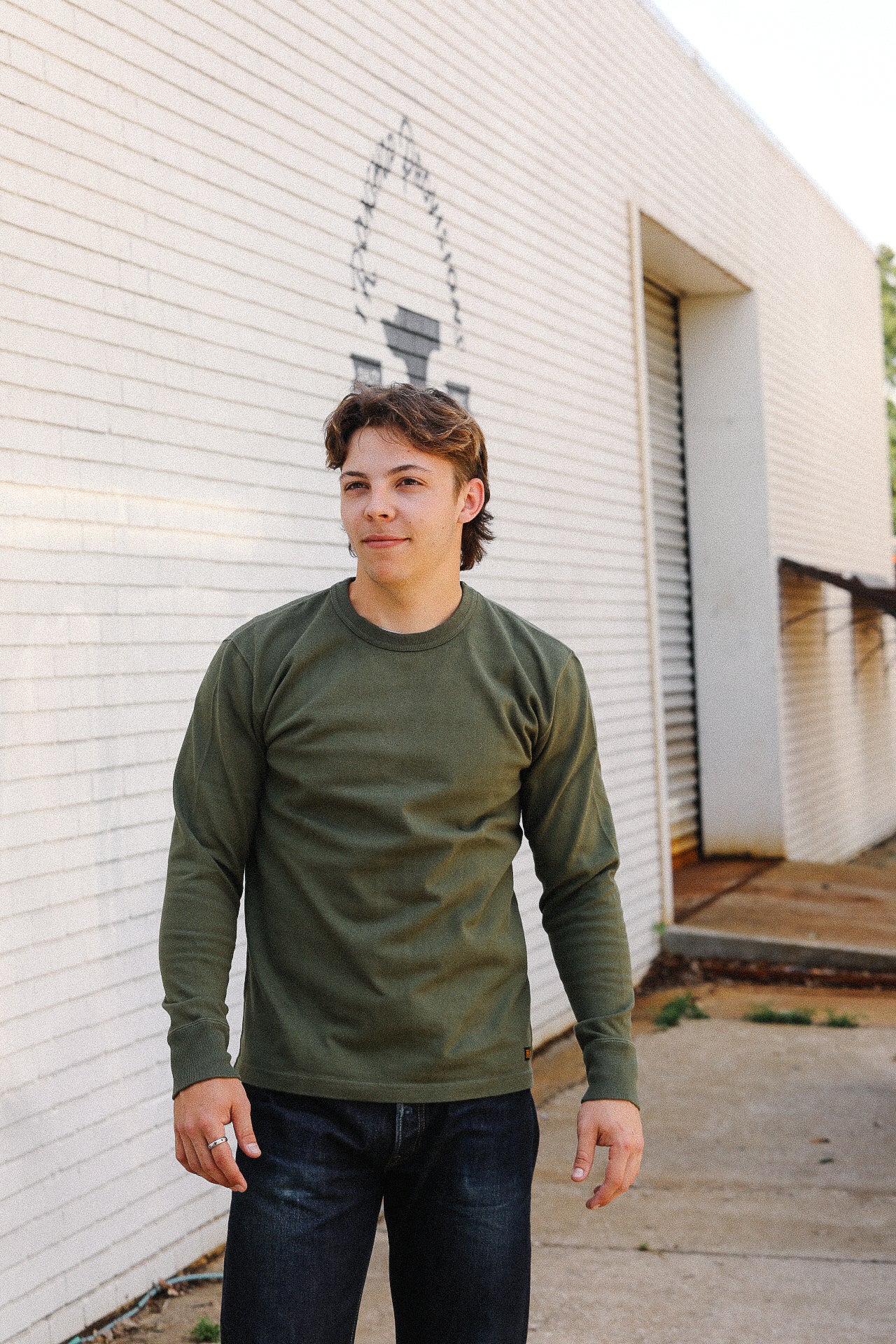 Young man in olive long-sleeve shirt and dark jeans standing outside an industrial white brick building