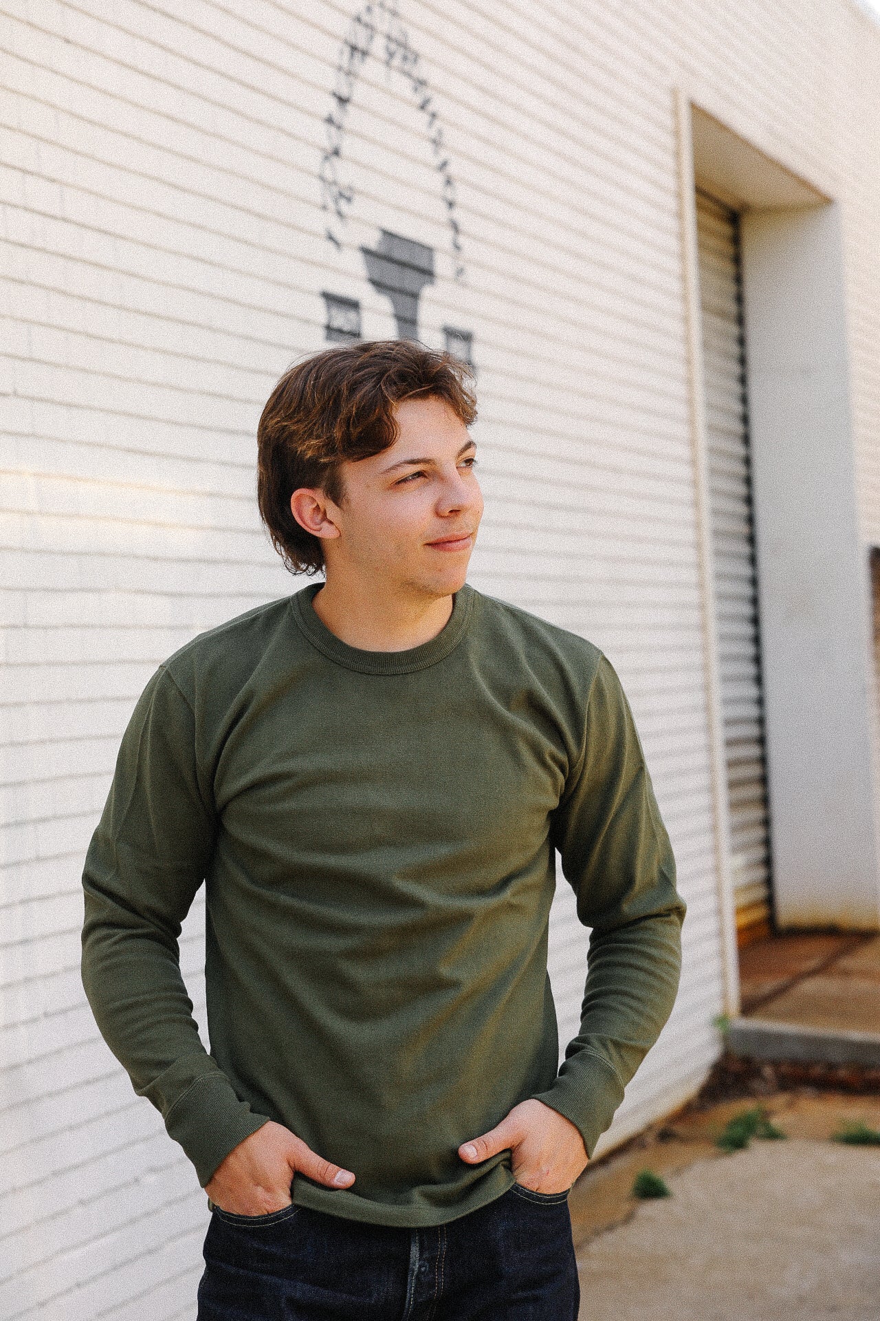 young man in olive green long sleeve shirt standing outdoors by white brick wall with blacksmith logo