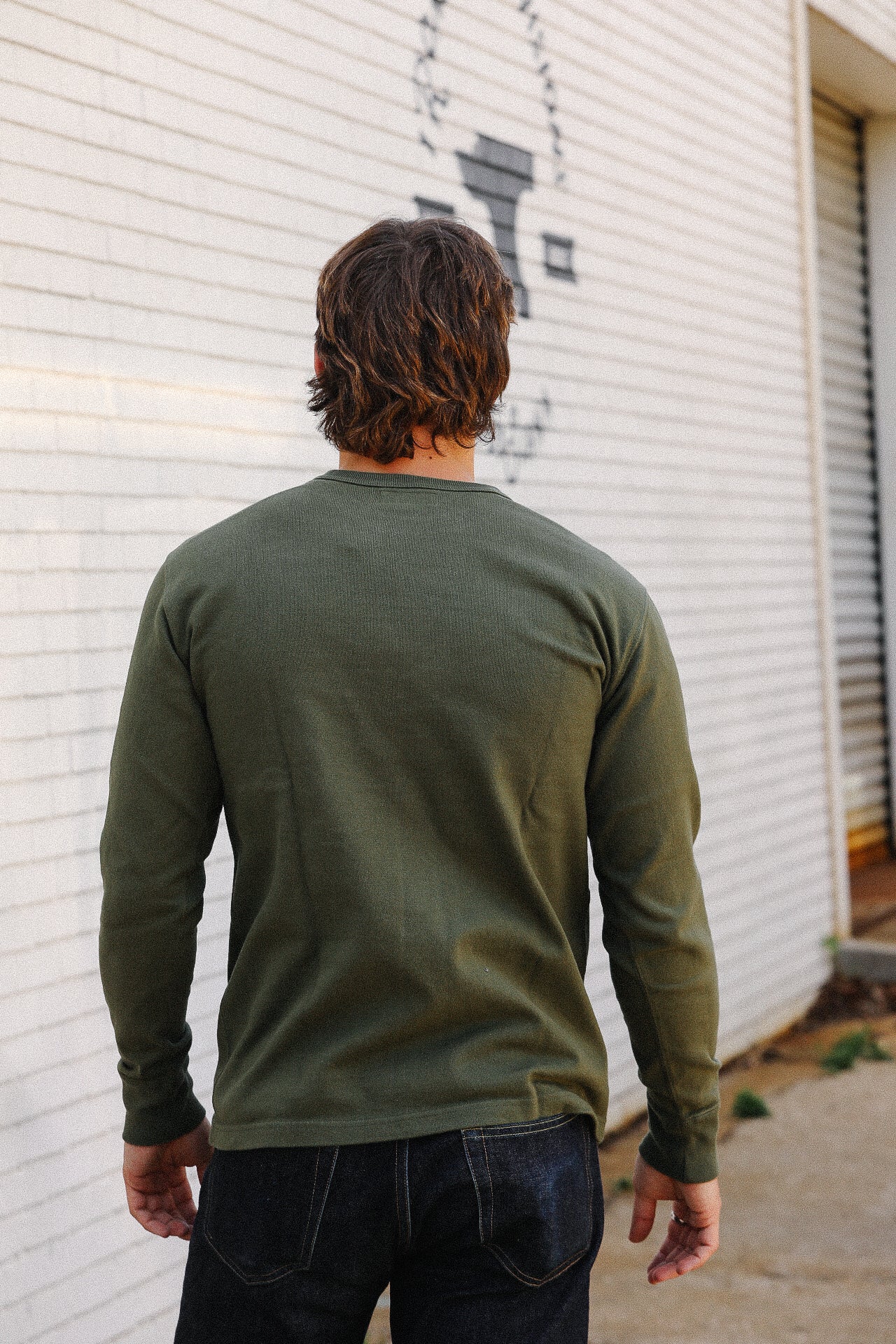 Man wearing olive green long sleeve shirt and dark jeans standing outside by white brick wall