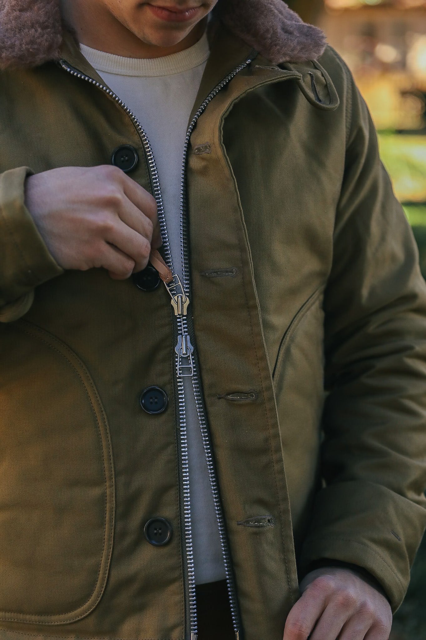 Close-up of a person wearing and zipping an olive green jacket with shearling collar and large black buttons