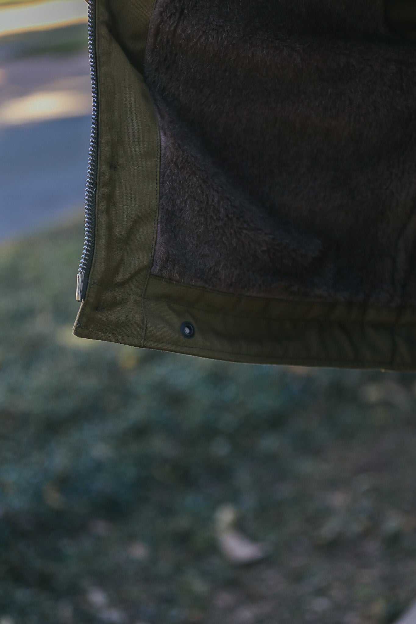 Close-up of olive green jacket hem with metal zipper and dark brown fleece lining, outdoor background