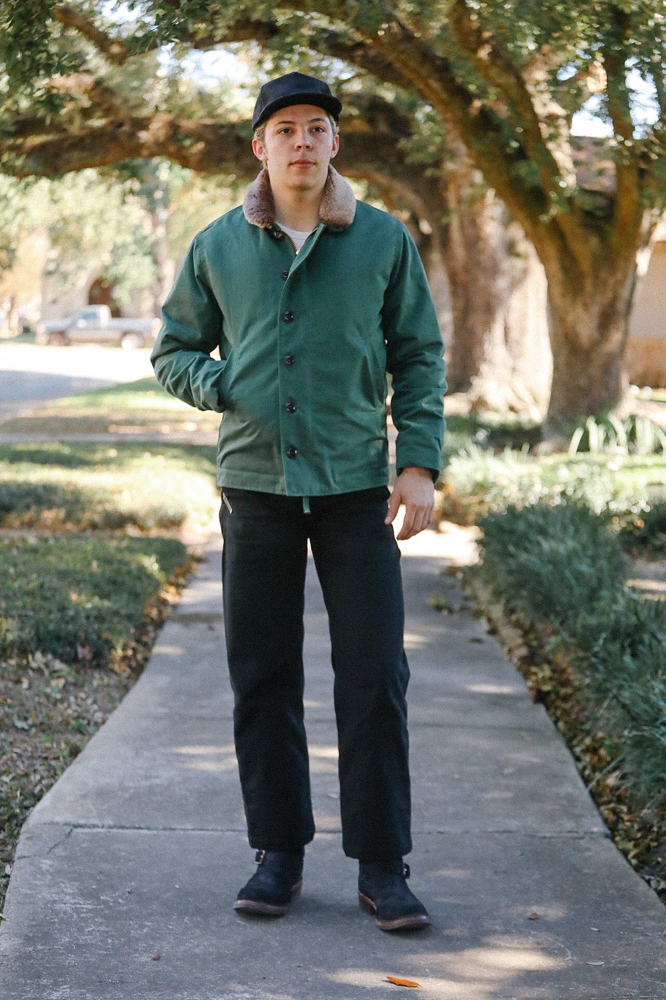 Man wearing green button-up jacket with shearling collar, black pants, cap, and boots standing on tree-lined sidewalk