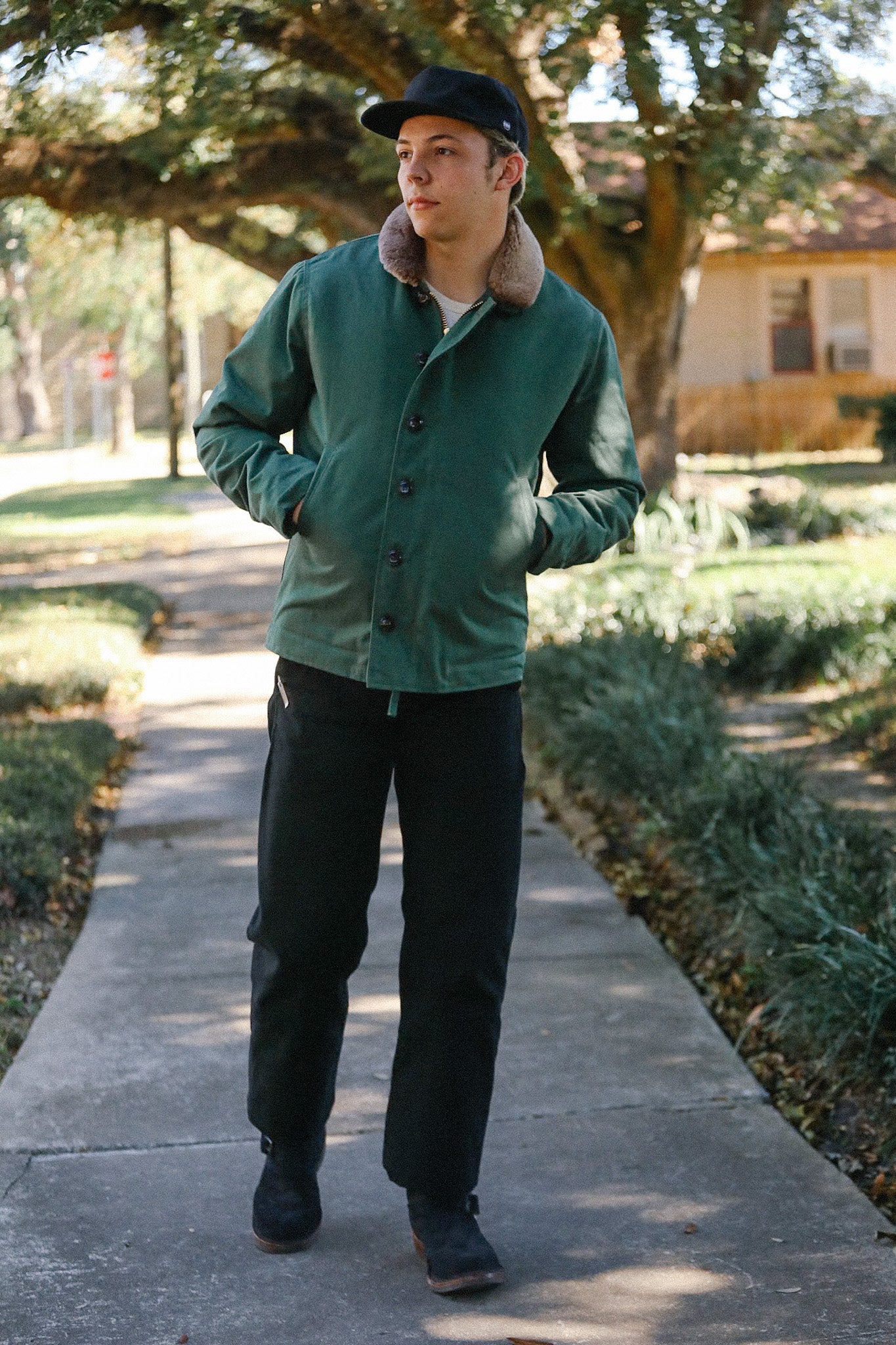 Man wearing green jacket with fur collar and black pants walking on sidewalk in park