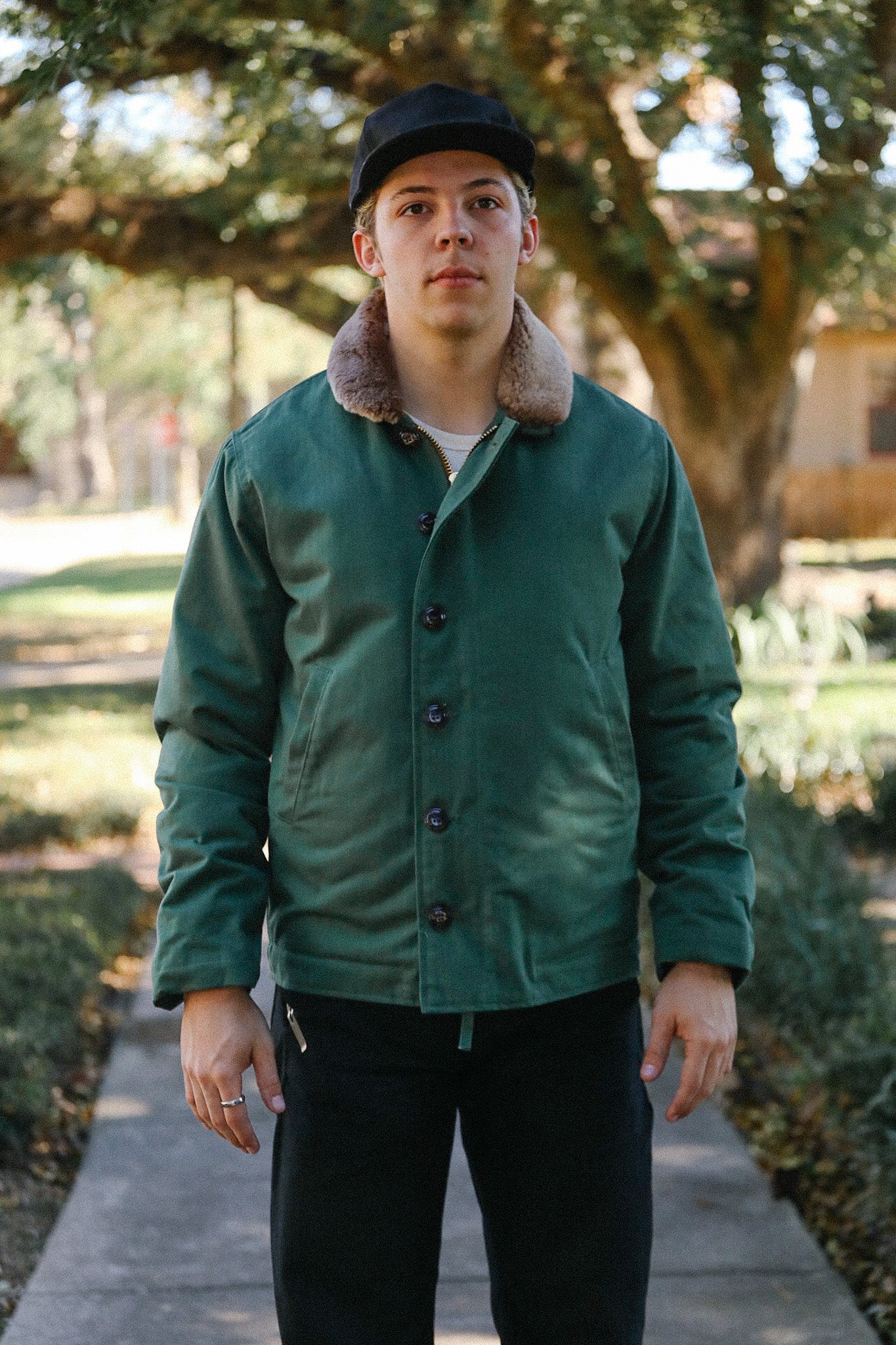 Young man wearing green jacket with fur collar and black cap standing outdoors on a tree-lined path