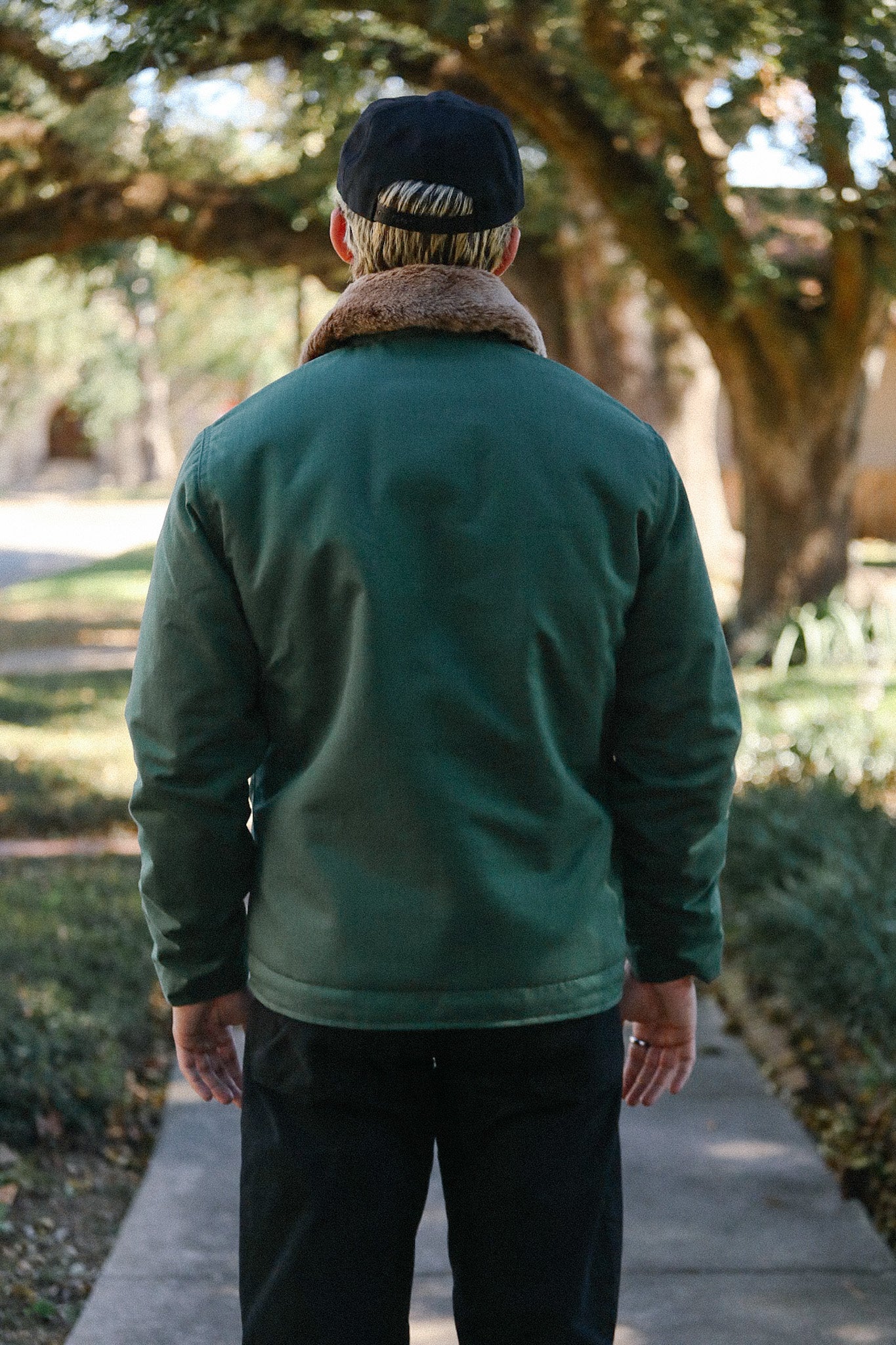Person wearing green jacket with brown fur collar and black cap standing on walkway outdoors