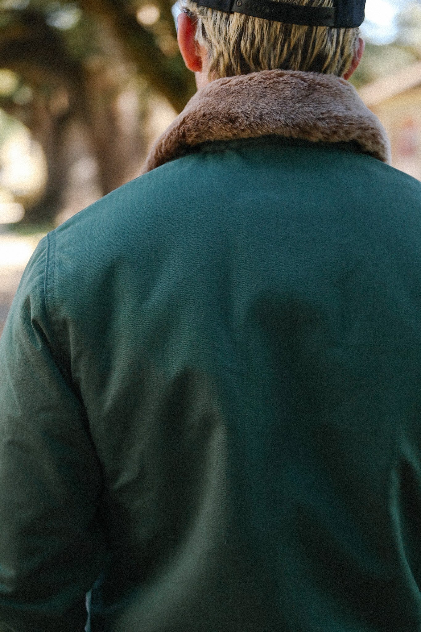 Back view of person wearing green jacket with brown fur collar and black cap, outdoors in daylight