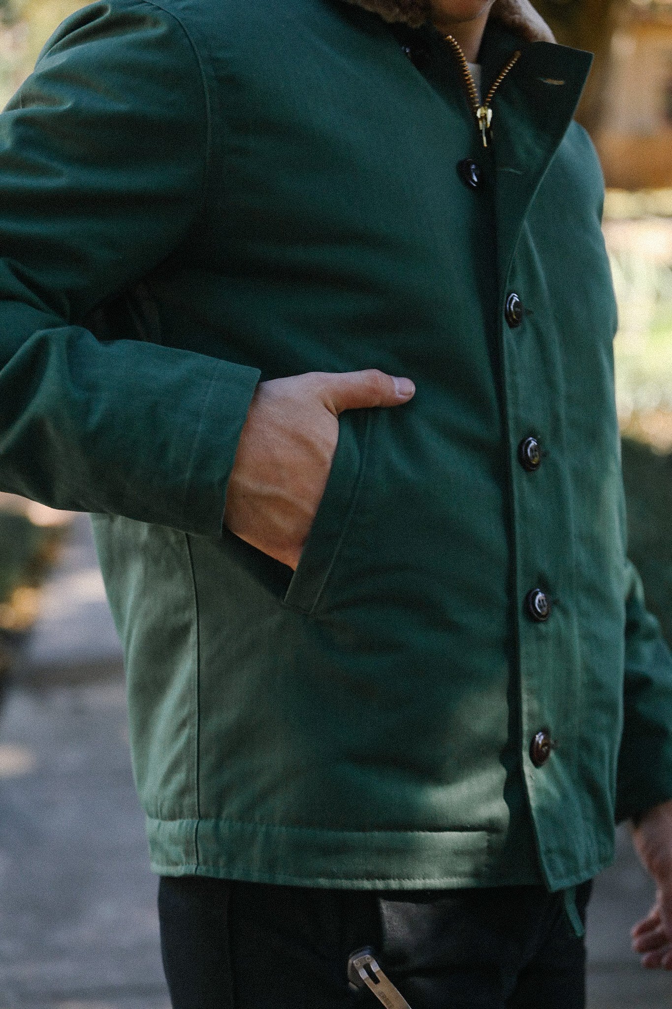 Close-up of a person wearing a green button-up jacket with hand in pocket, outdoor setting
