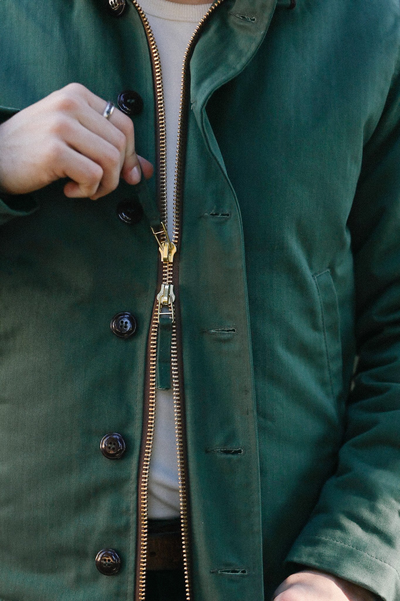 Close-up of person unzipping a green jacket with brass zipper and black buttons, wearing a silver ring