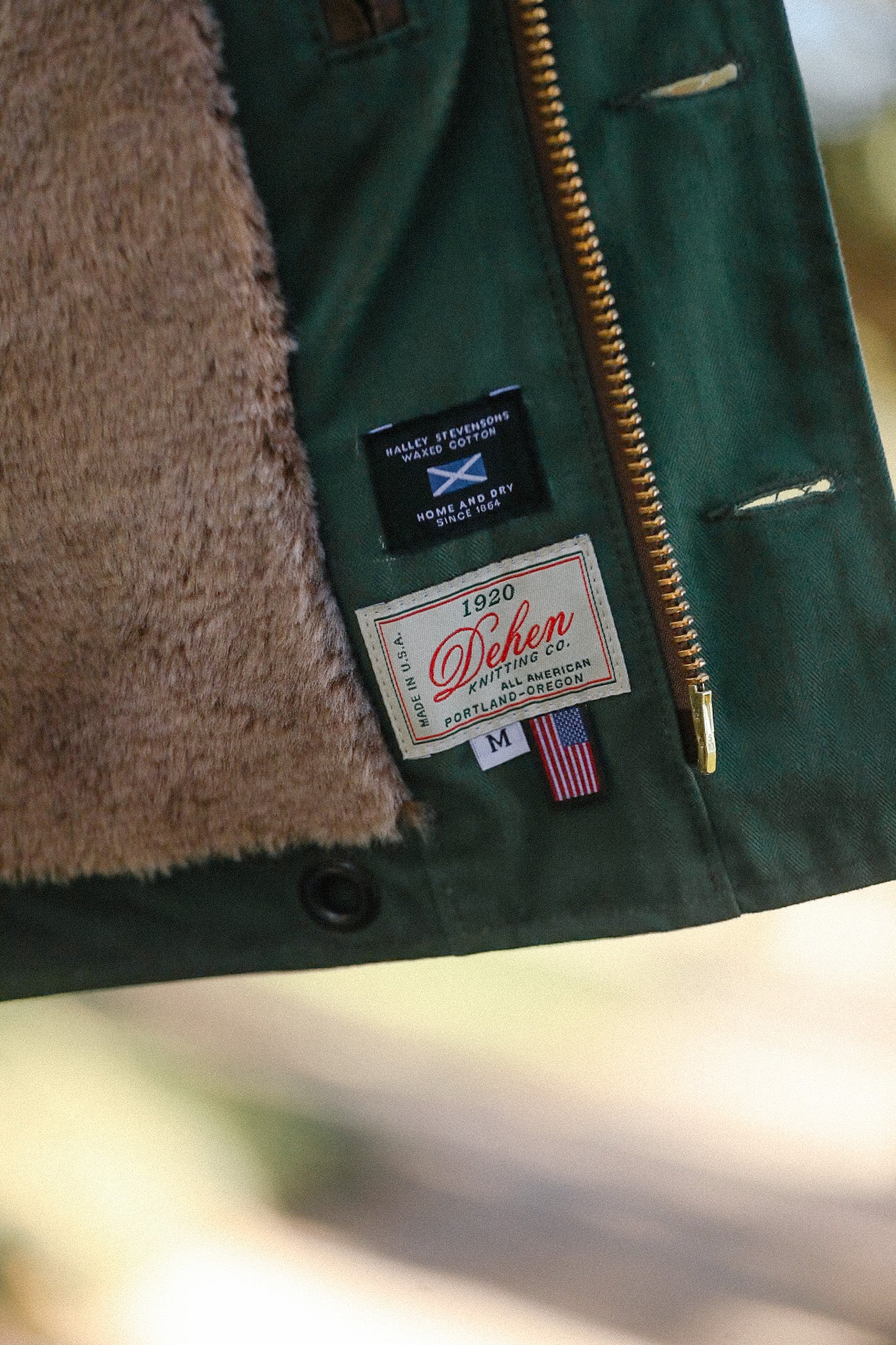 Close-up of lined green jacket showing Dehen Knitting Co. label, waxed cotton tag, and zipper detail