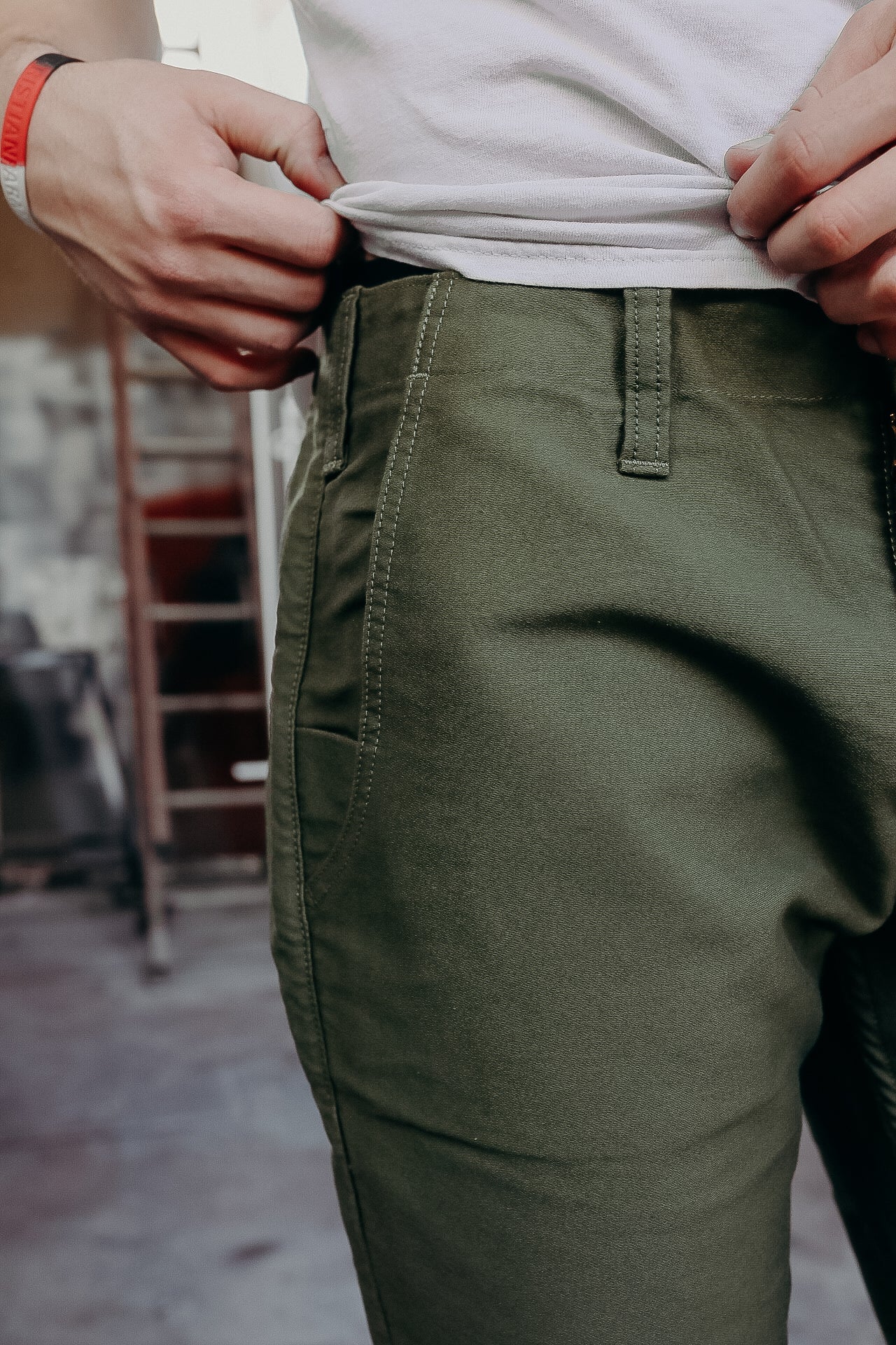 Close-up of person wearing olive green durable work pants and adjusting white shirt in workshop