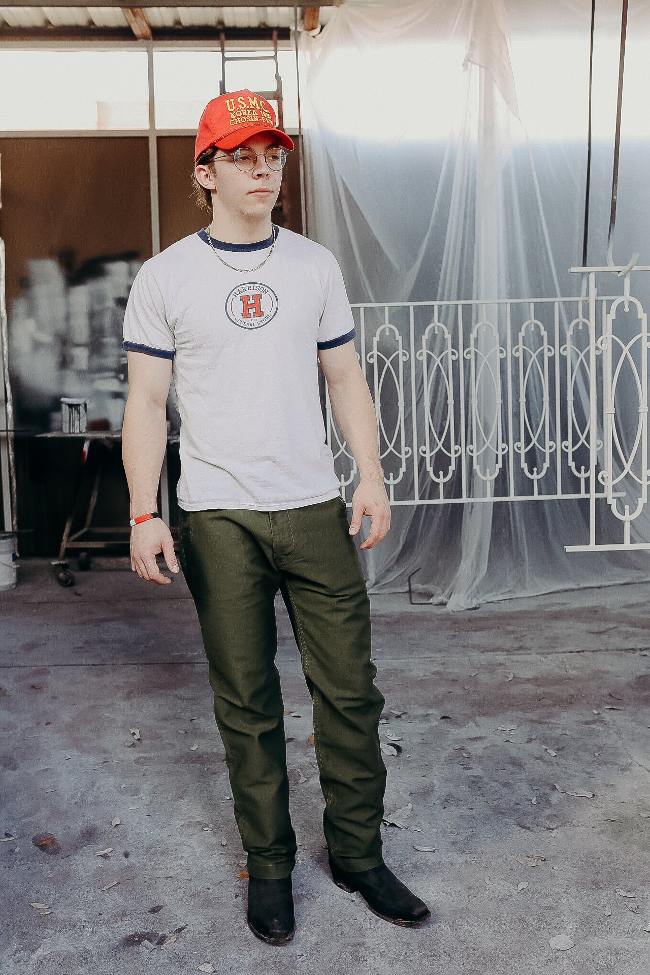 Man wearing vintage white Harrison General Store t-shirt, olive green pants, black boots, and red USMC cap indoors