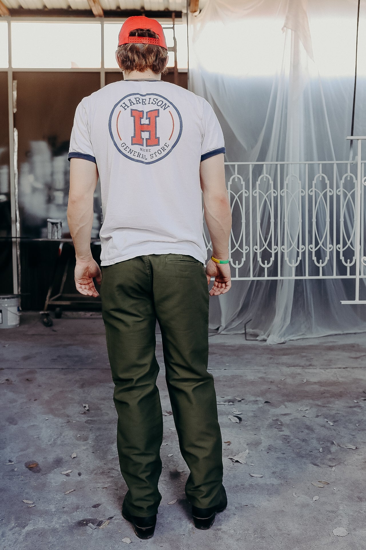 Man wearing white Harrison General Store t-shirt and olive green pants standing indoors near iron railing