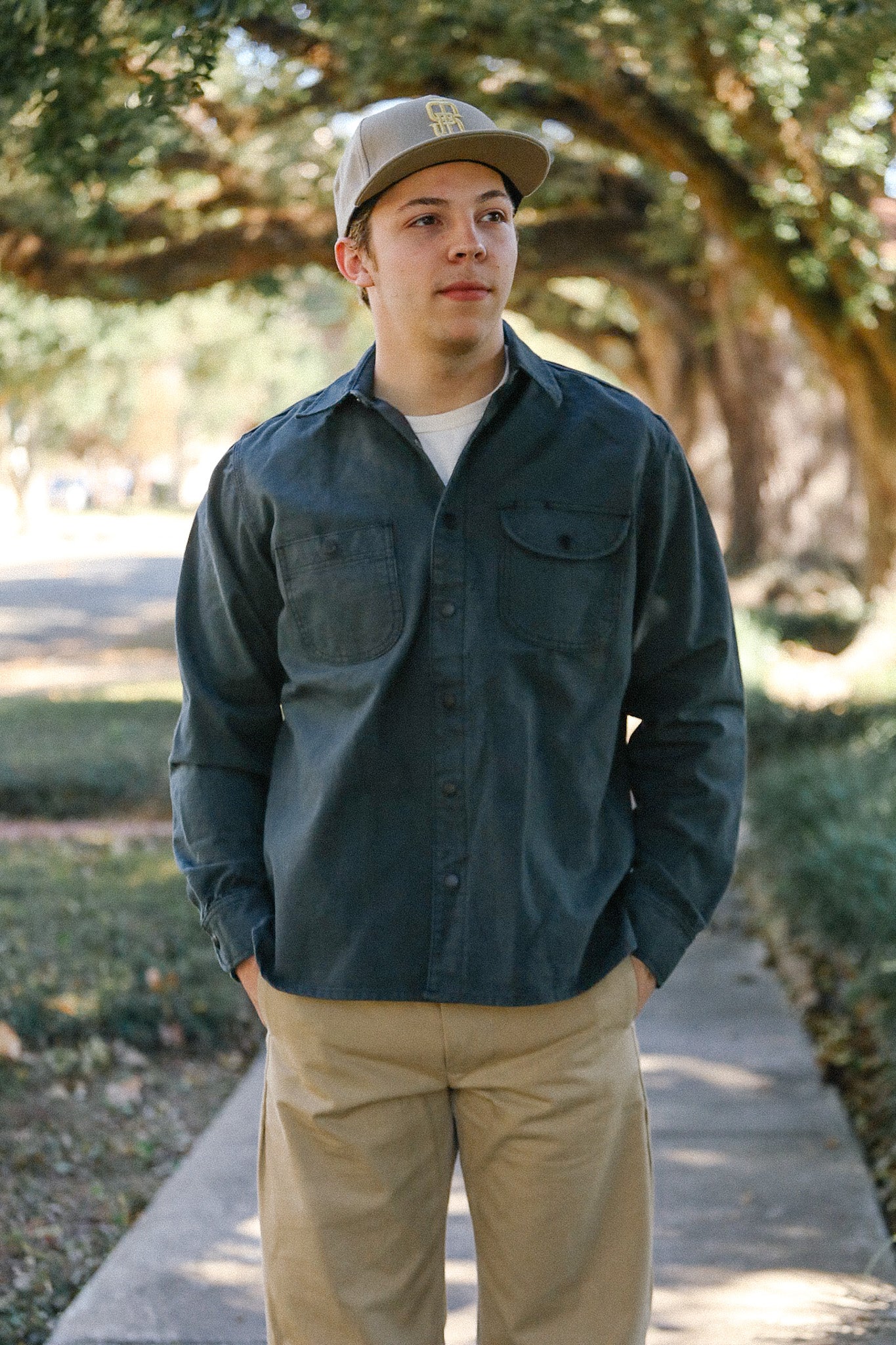 Young man wearing a dark button-up shirt, khaki pants, and beige cap standing outdoors on a shaded pathway