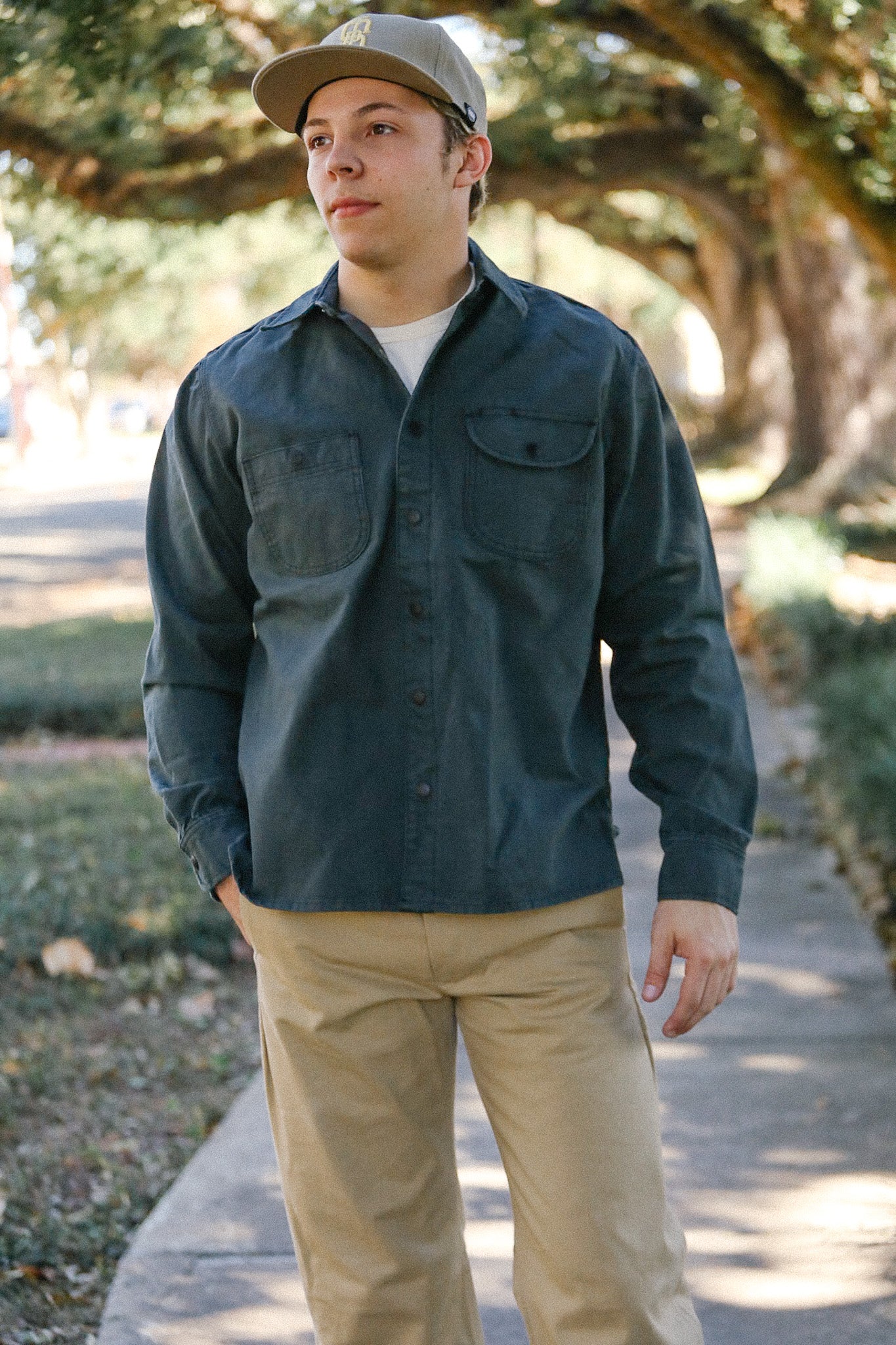 Man wearing a dark button-up shirt, khaki pants, and beige cap walking on a tree-lined sidewalk