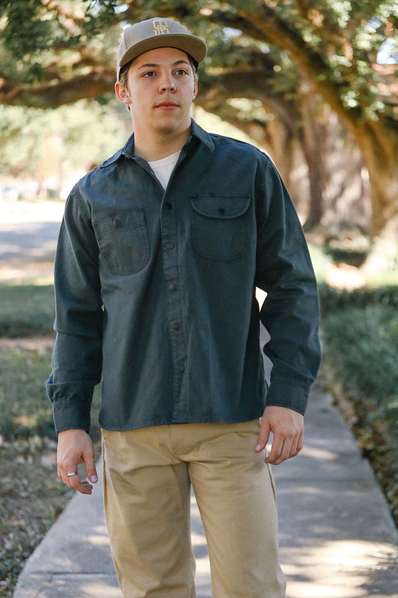 Young man wearing a dark green work shirt and tan pants walking outdoors on a tree-lined path