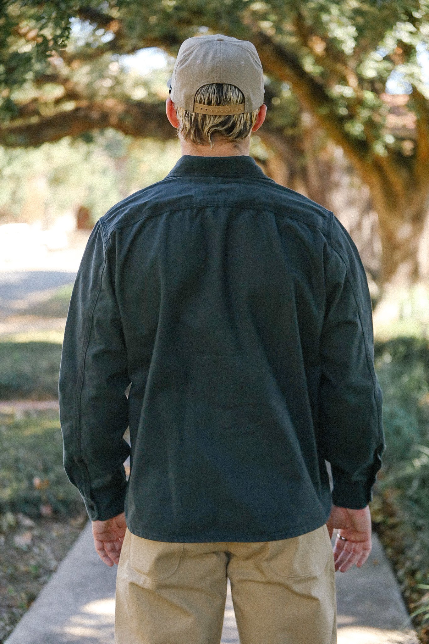 Man wearing a dark long-sleeve shirt and beige pants, standing on a pathway under trees outdoors