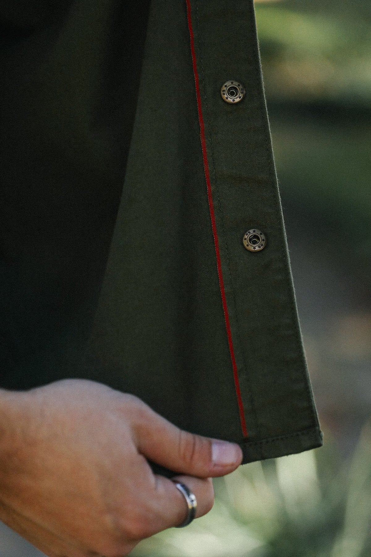 Close-up of hand holding olive green garment hem with metal snap buttons and red stitching detail
