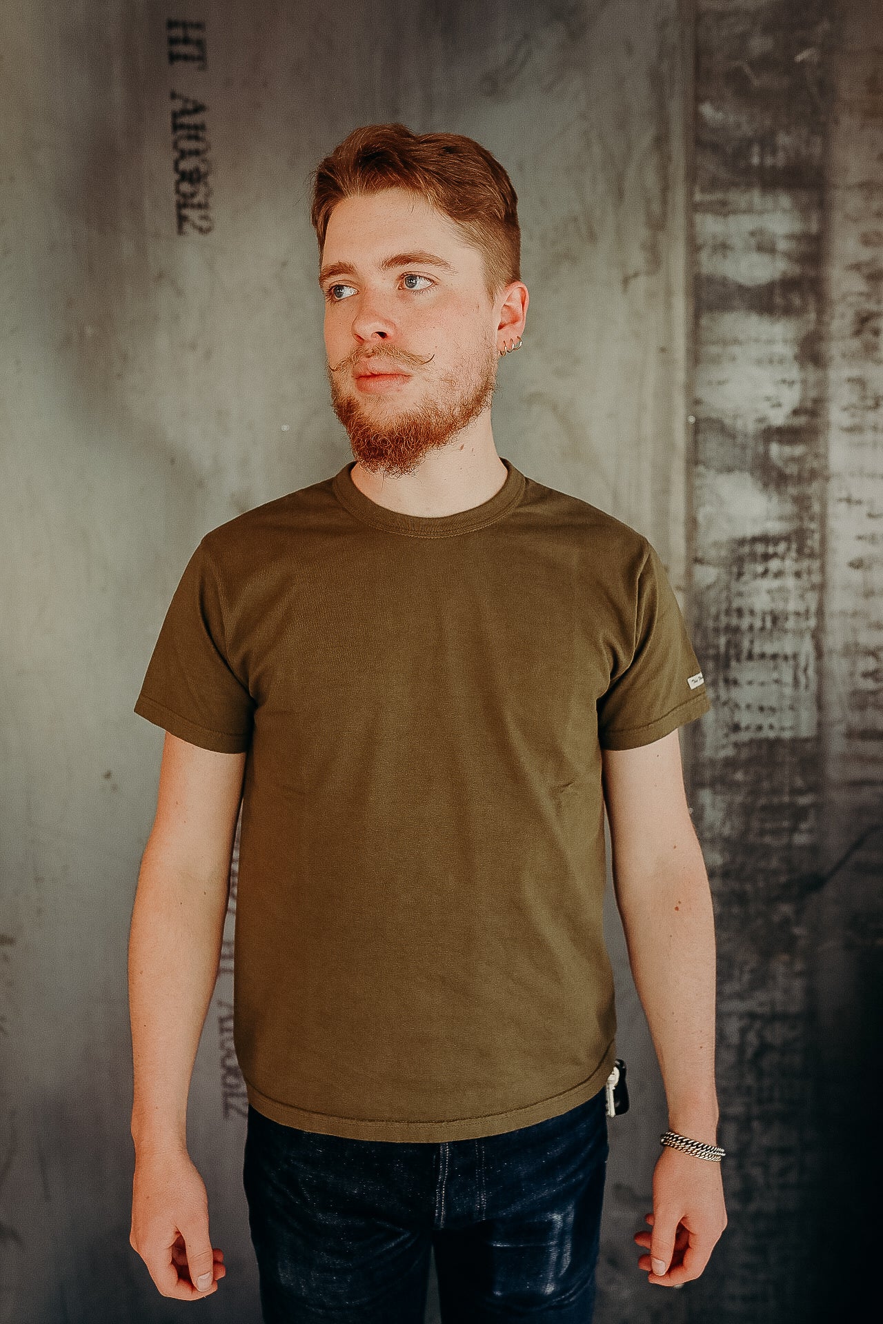 Man with styled beard wearing olive green t-shirt and dark jeans against industrial concrete background