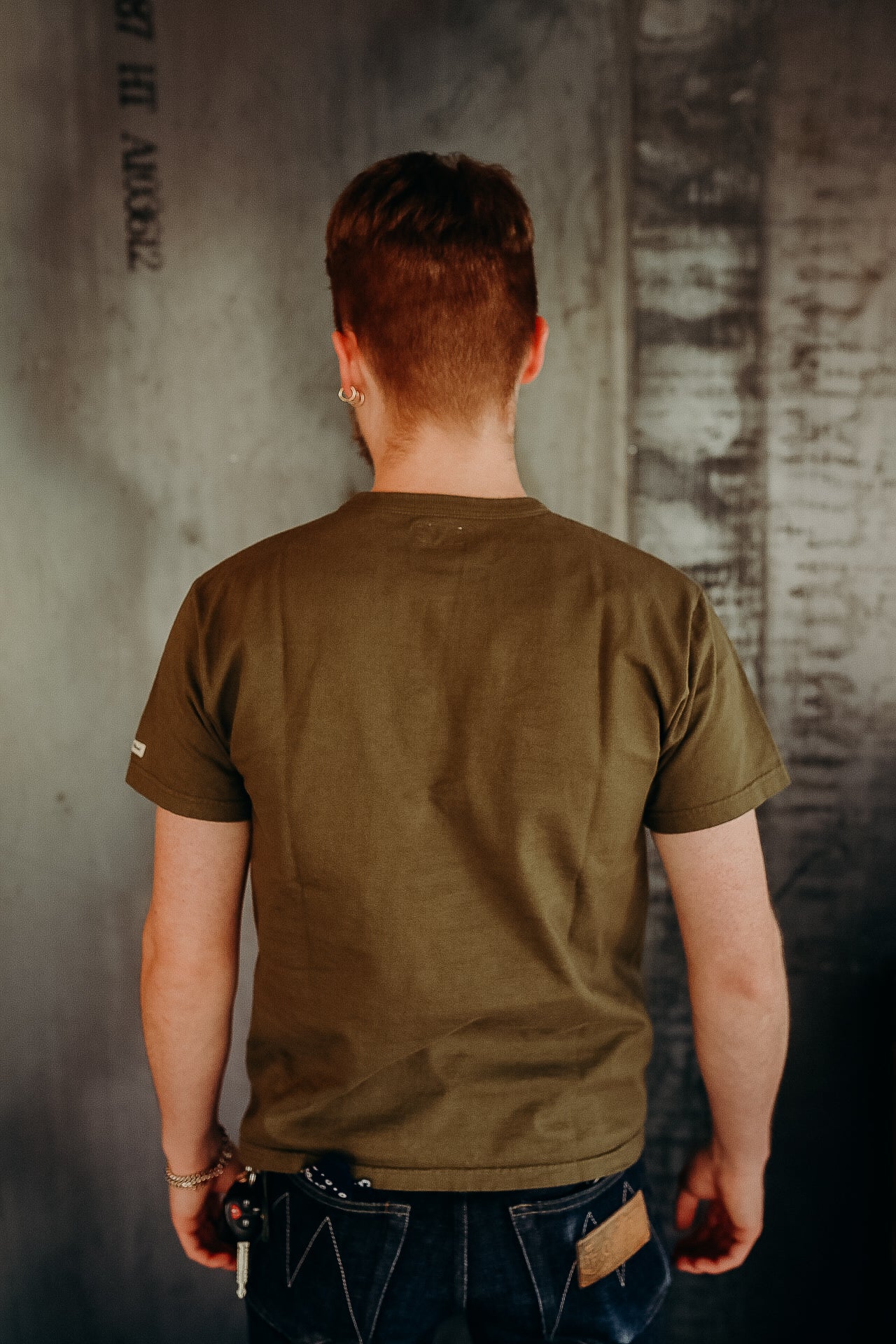 Man wearing olive green short-sleeve t-shirt and dark denim jeans standing against industrial wall