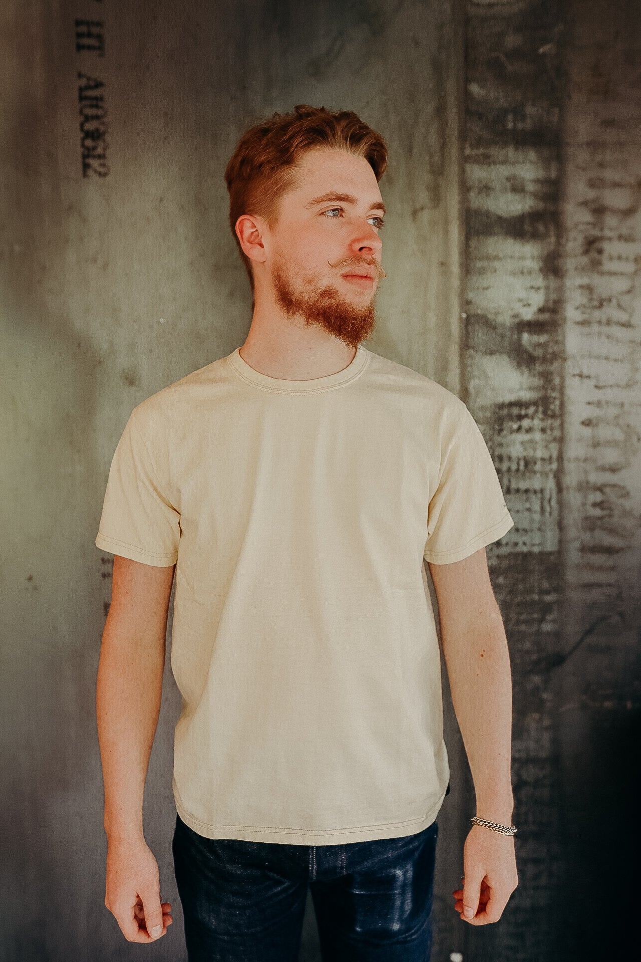 Young man with a beard wearing a beige t-shirt and dark jeans standing against a textured industrial wall