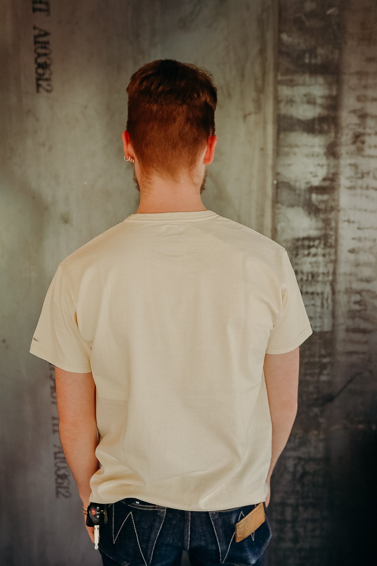 Back view of man wearing beige t-shirt and dark jeans standing against textured industrial wall