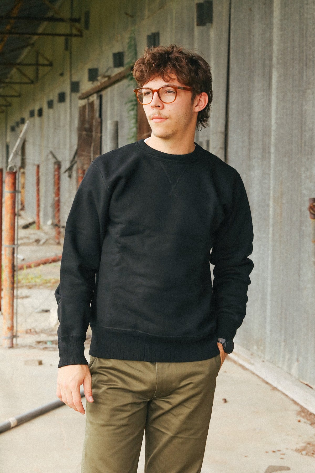 A man wearing a black crewneck sweatshirt and olive green pants, standing in front of a gray concrete wall.