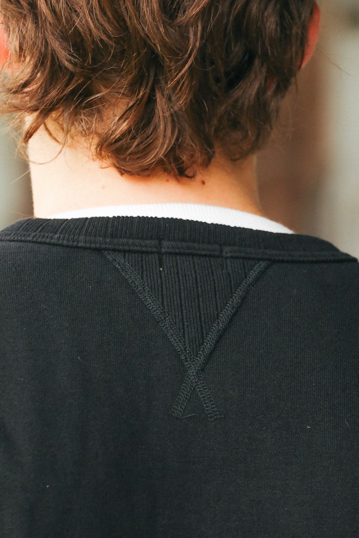 Close-up back view of a person wearing a black crewneck sweatshirt with a visible Yoke Stitch detail on the upper back, layered over a white undershirt.