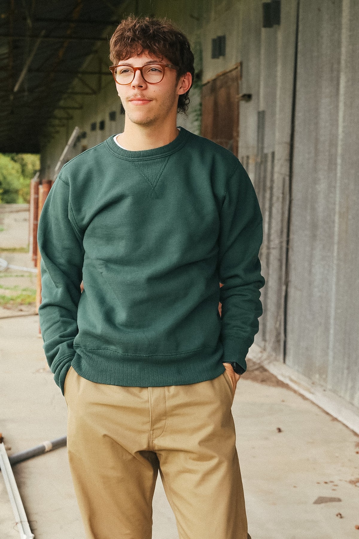 A man wearing a dark green crewneck sweatshirt and tan pants, standing in front of a rustic concrete wall.