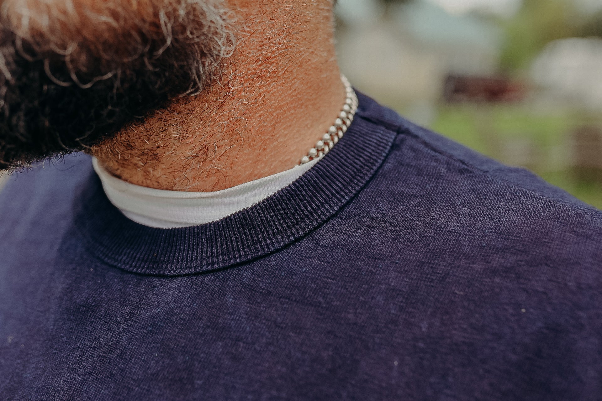 Close-up of a man with a beard wearing a silver chain necklace and navy blue crewneck shirt