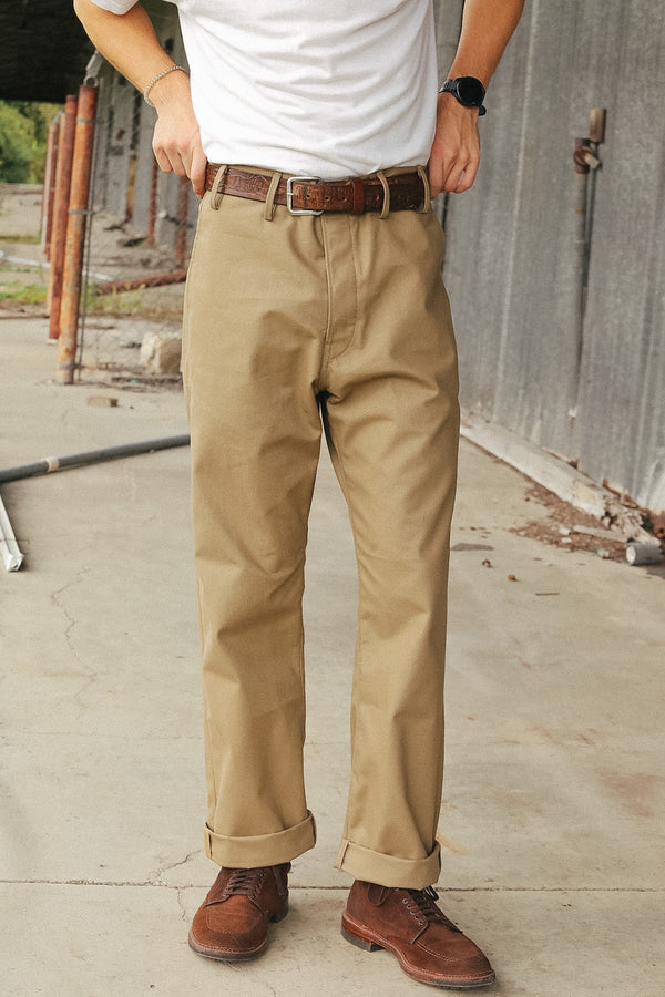 A person wearing khaki chinos with a brown leather belt and brown leather shoes, standing outdoors in front of a gray metal wall.